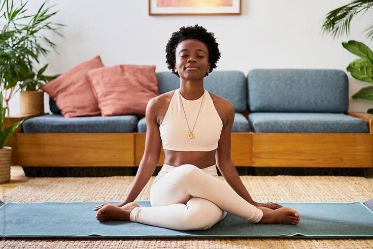 A young Black woman wearing a cream-colored sports bra and leggings sits on a blue yoga mat with her legs crossed in a yoga pose. Her eyes are closed in meditation, and she wears a calm expression. She is in a bright, cozy living room featuring a blue sofa with pink pillows, a woven rug, and lush potted plants in the background.