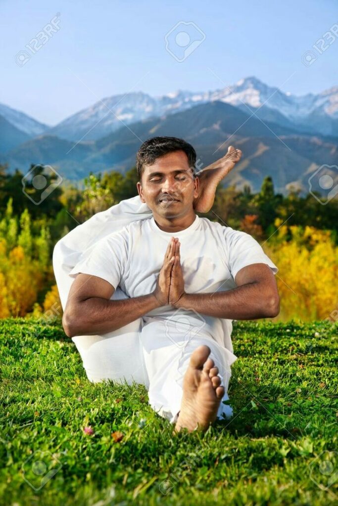 A man dressed in white clothing sitting on vibrant green grass performing an advanced yoga pose, with one leg wrapped behind his neck and his hands pressed together in a prayer position. His eyes are closed in meditation, with a scenic background of autumn trees, majestic mountains, and a clear blue sky.